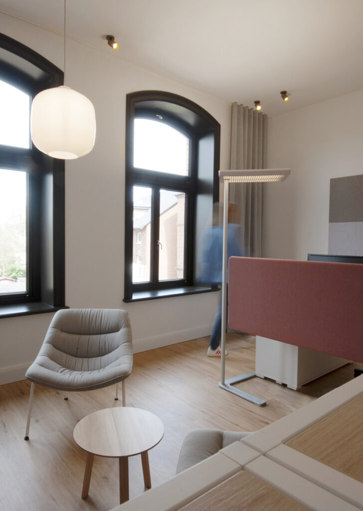Büroraum mit Schreibtisch und roter Tischtrennwand neben einer Stehleuchte und einem Loungeberich mit Sesseln und Pendelleuchte